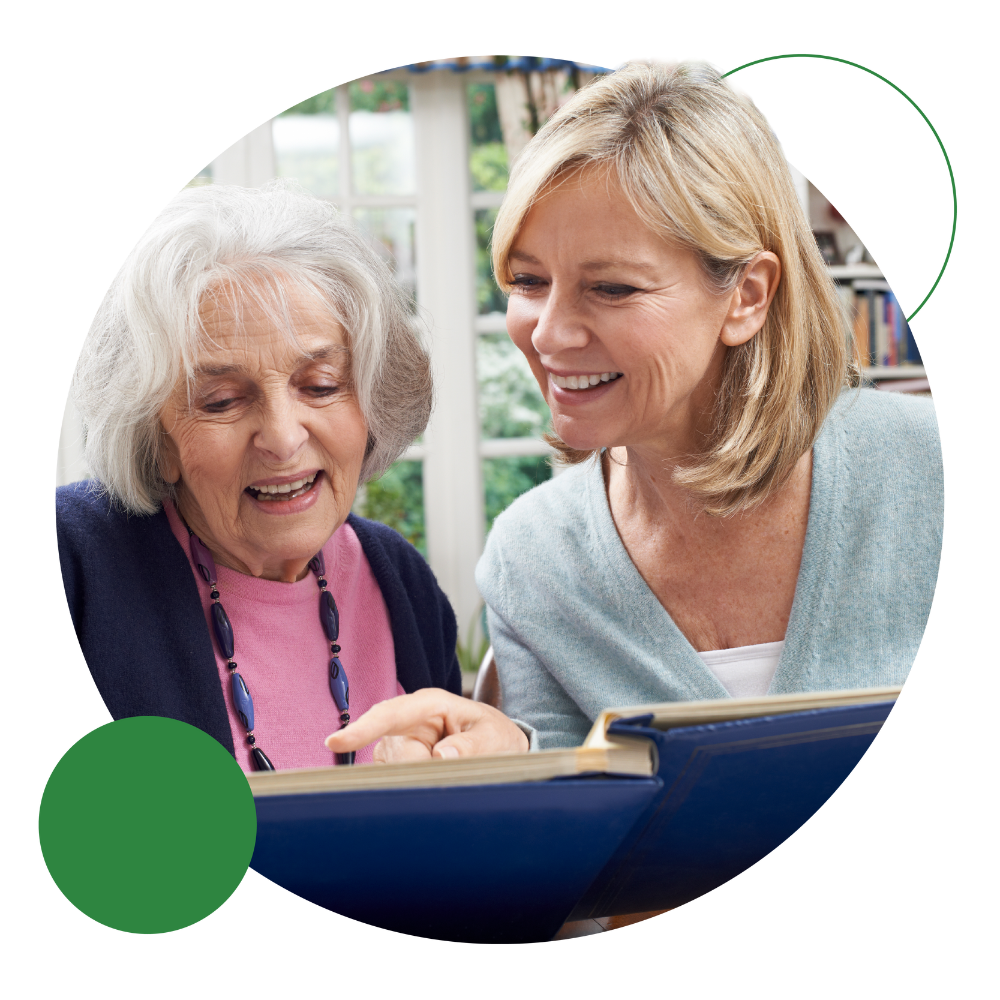 Lady-Reading-Book-In-Care-Home (1) 5 Older person in a a care home setting reading a large book with a visitor beside them