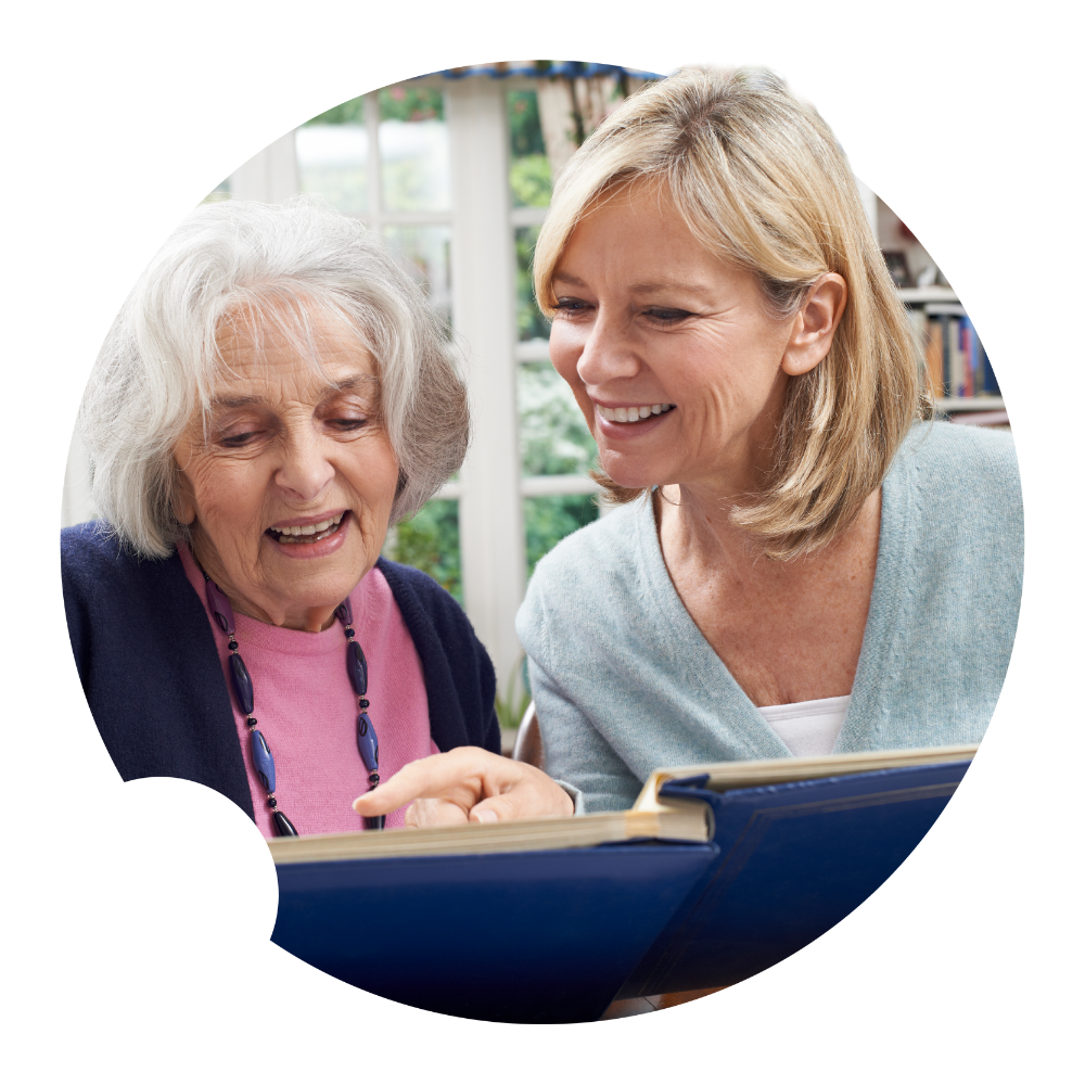 Older person in a a care home setting reading a large book with a visitor beside them