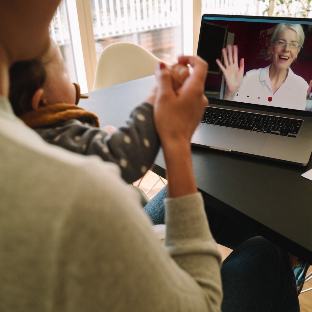 Parent holding baby during a video call with Liaison Care expert on a laptop at a table.
