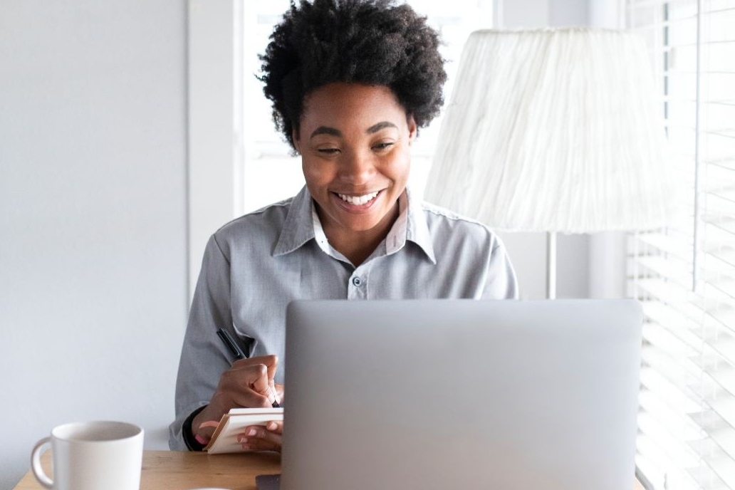 Building A Stronger Workforce With England’s First Postgraduate Diploma In Continuing Healthcare 1 A woman sitting at a desk using her laptop and writing notes, participating in continuing healthcare postgraduate diploma